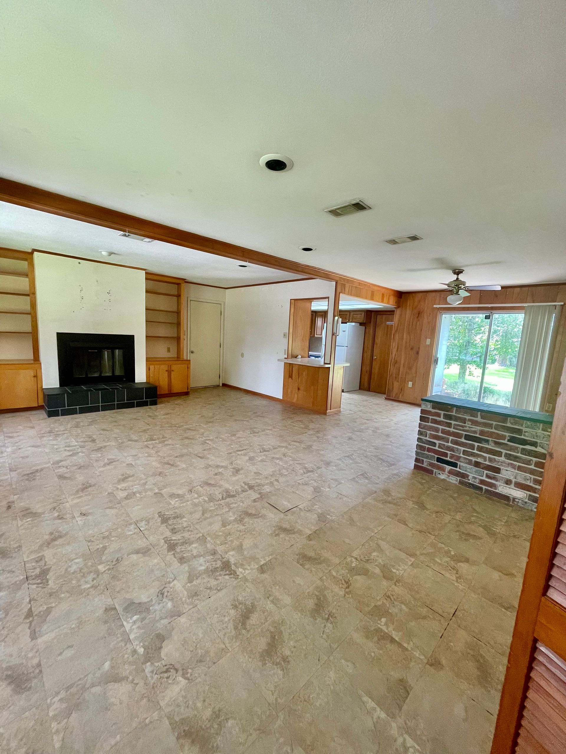 Empty living room with fireplace and exposed beams. Brown paneling, beige flooring, and a brick accent.
