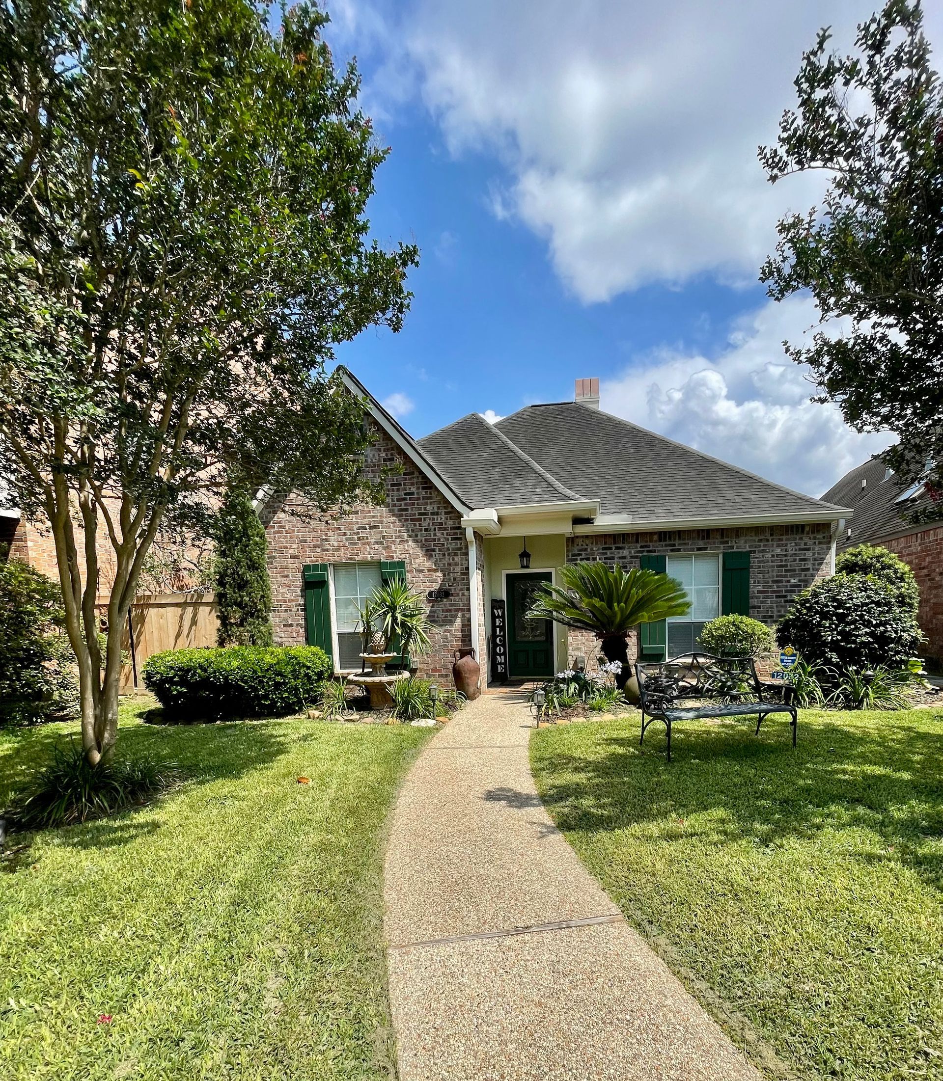 Brick house with green shutters, a stone path, and lush landscaping under a cloudy sky.