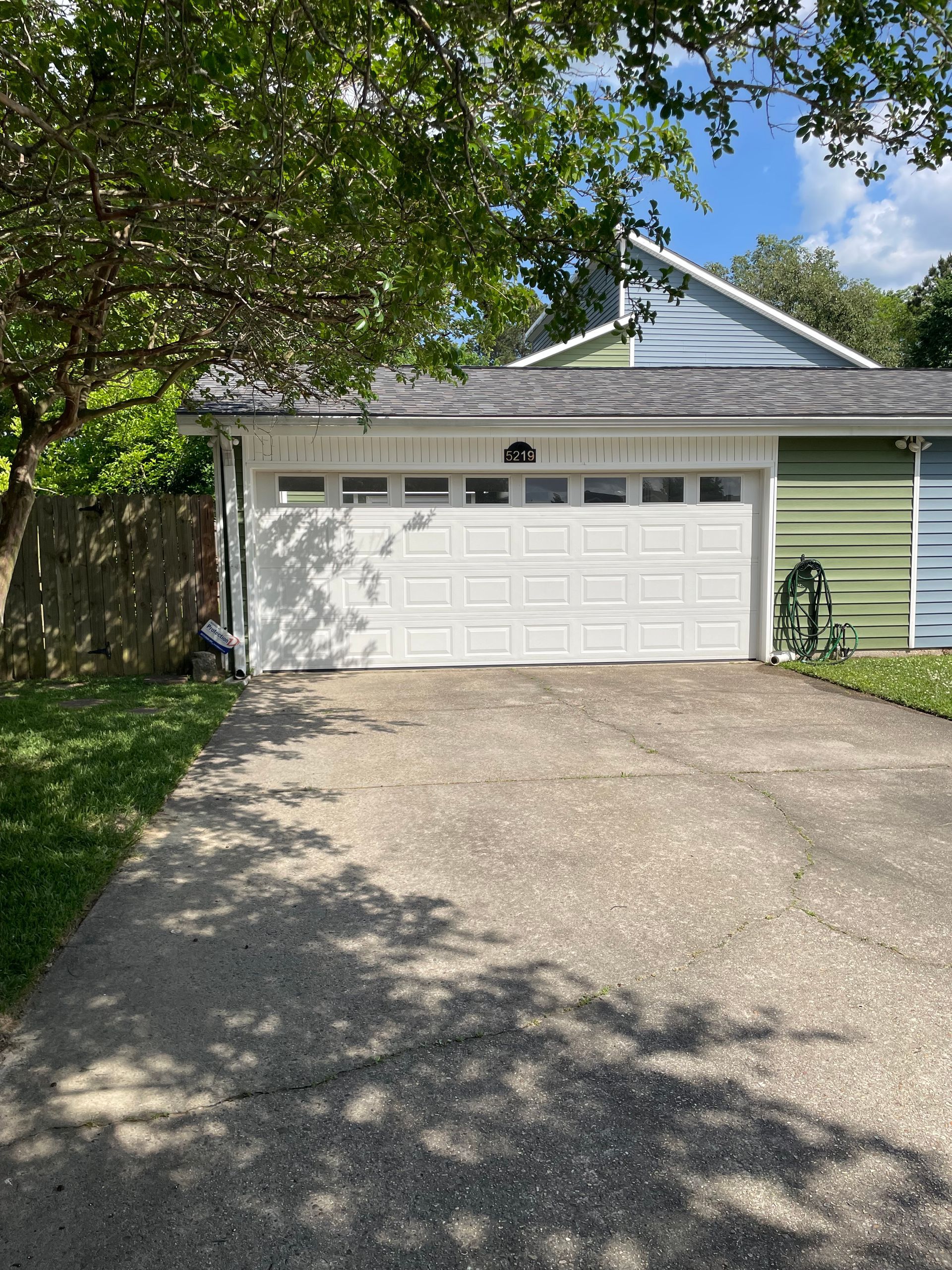 Suburban house with white garage door, green siding, and a tree-shaded driveway