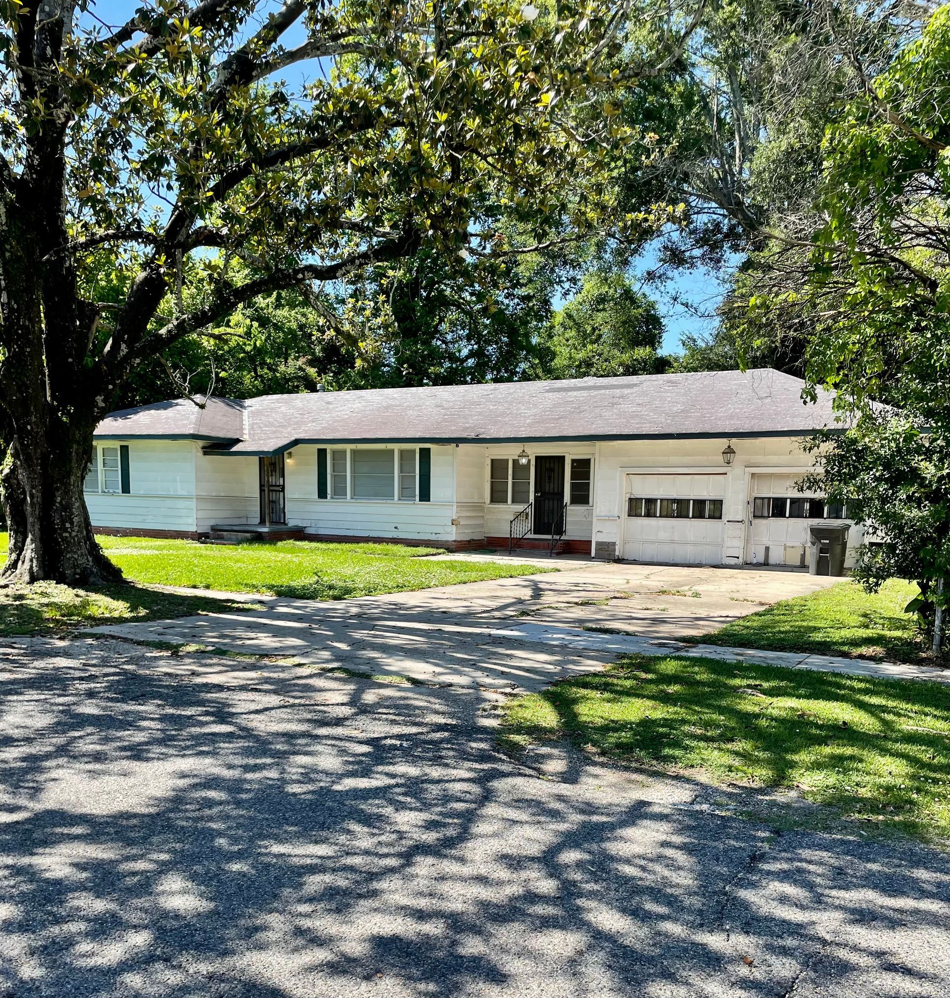 Single-story white house with a garage, set behind large trees and a gravel driveway on a sunny day
