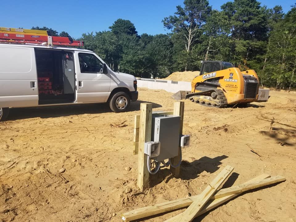 Construction site: Van, skid steer, electrical box on posts, and foundation. Sandy ground under a blue sky.