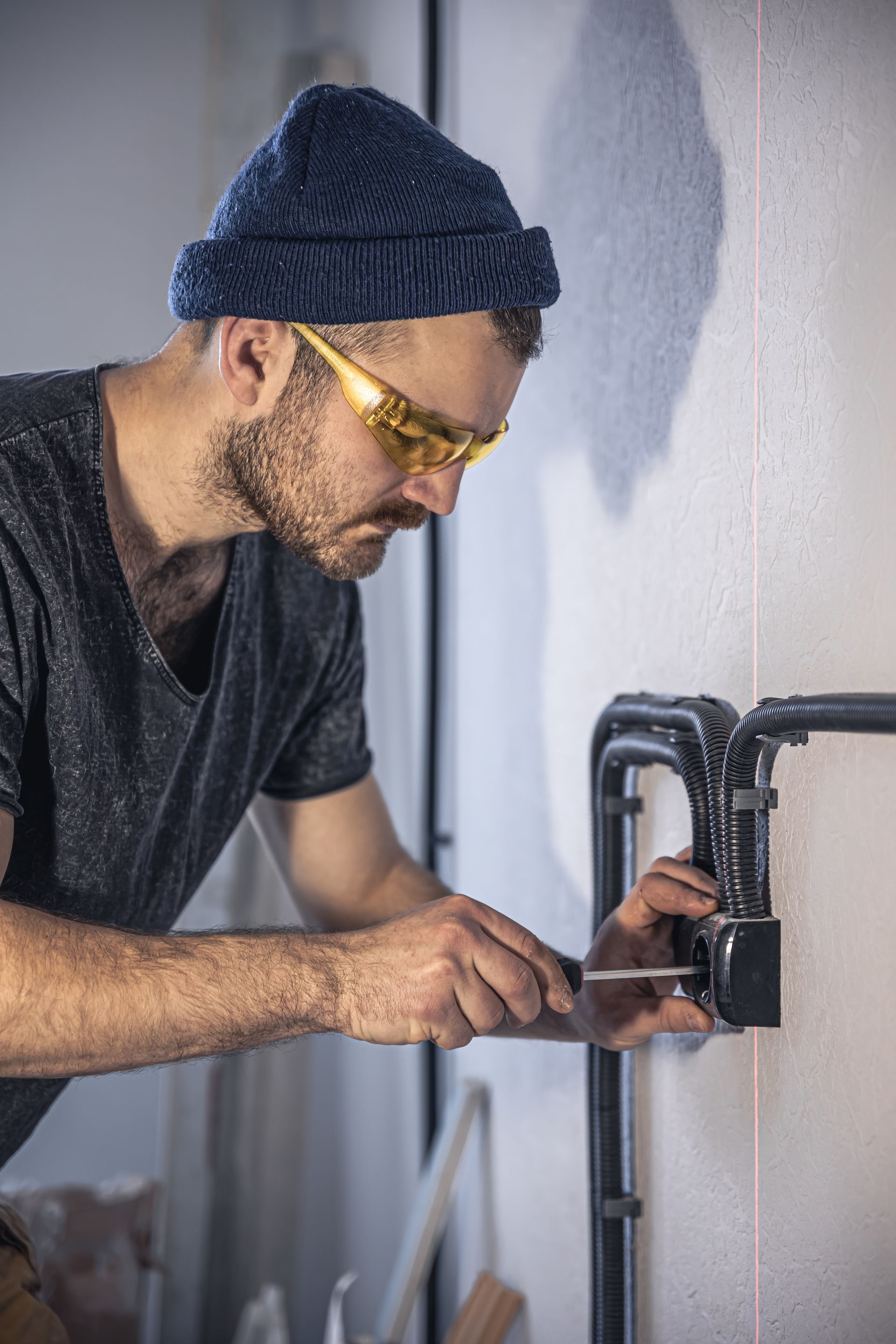 Man in beanie and safety glasses using a screwdriver on black pipes attached to a white wall.