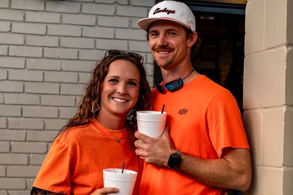 Couple in orange shirts smiling, holding drinks, near brick wall.