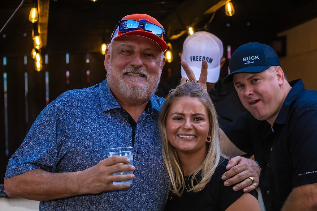 Four people smiling at camera. Man in hat with drink, woman, man behind woman with peace sign, man on right. Outdoors, night.