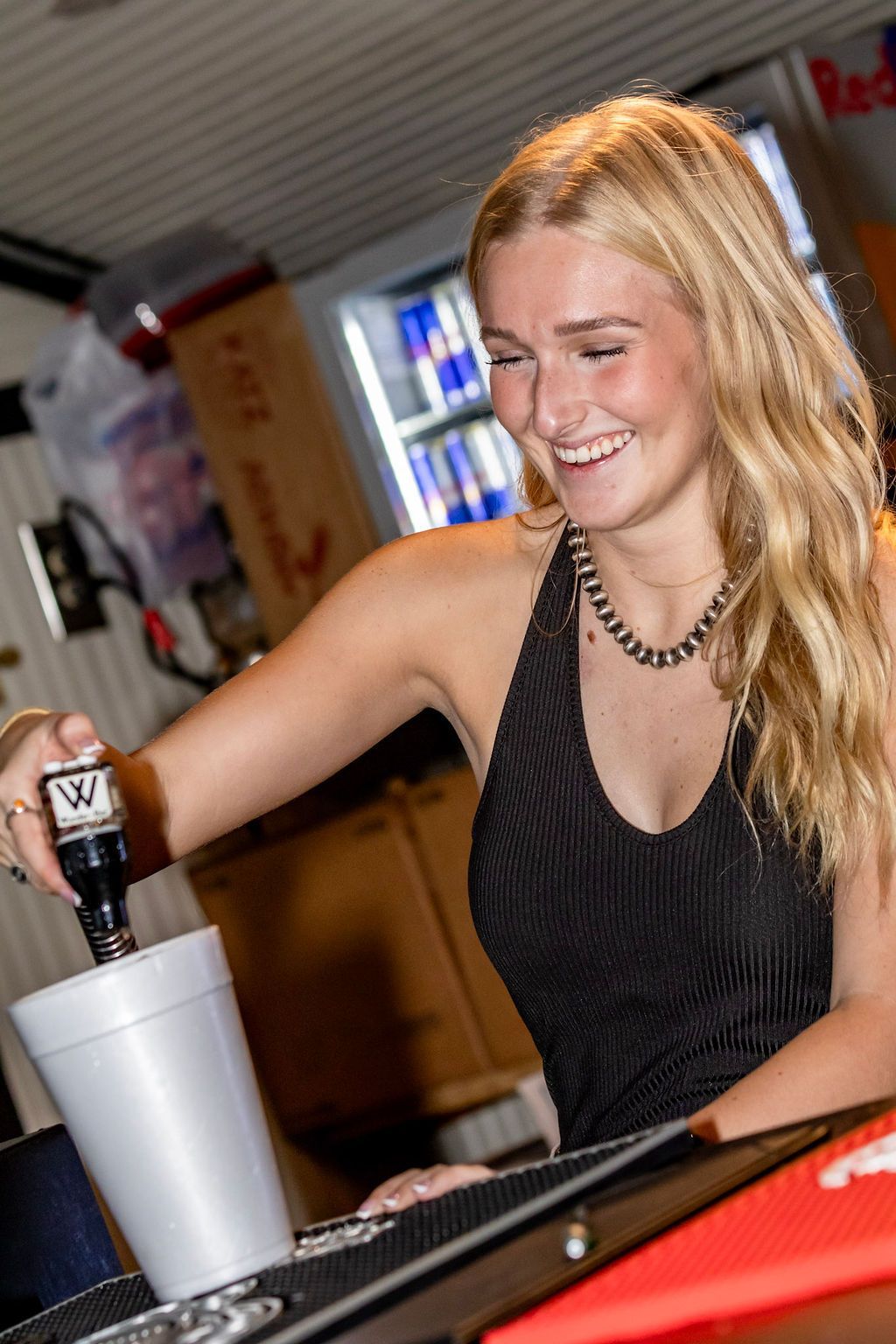 Blonde woman pours a beverage from a bottle into a white cup, smiling. Bar setting with Red Bull display.
