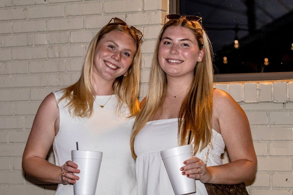 Two women smiling, holding drinks, wearing white, standing against a brick wall outside.