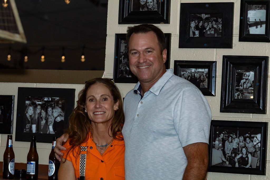 Woman in orange and man in blue smile, pose together near framed photos on a wall.