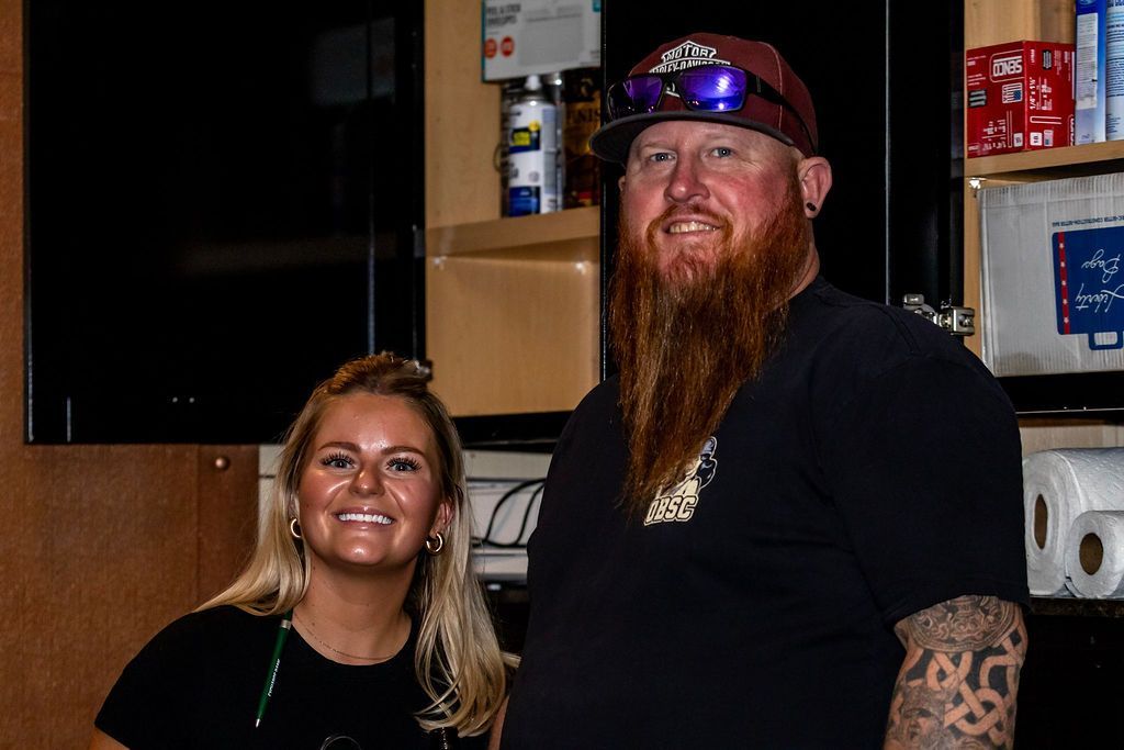 Two people smiling, indoors. A woman with blonde hair and a man with a red beard stand next to each other.