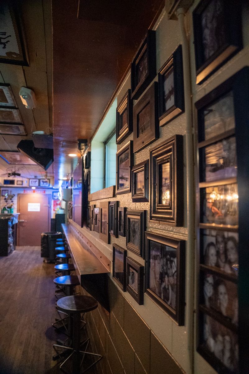 Bar interior with wood paneling, framed photos on the wall, and stools along a countertop.