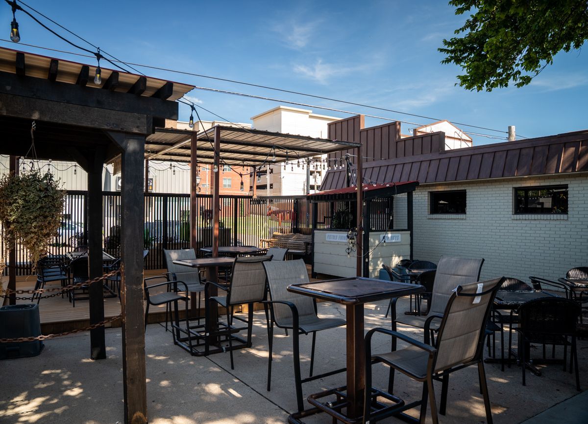 Outdoor patio with tables and chairs under a pergola, strung with lights, sunny day.
