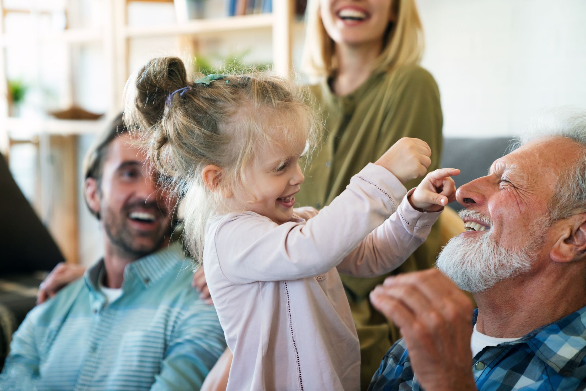 Girl playfully pokes a smiling elderly man’s nose. Family laughs in a cozy living room.