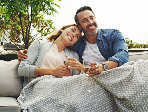 Couple snuggled under a blanket outdoors, smiling while holding glasses of wine.