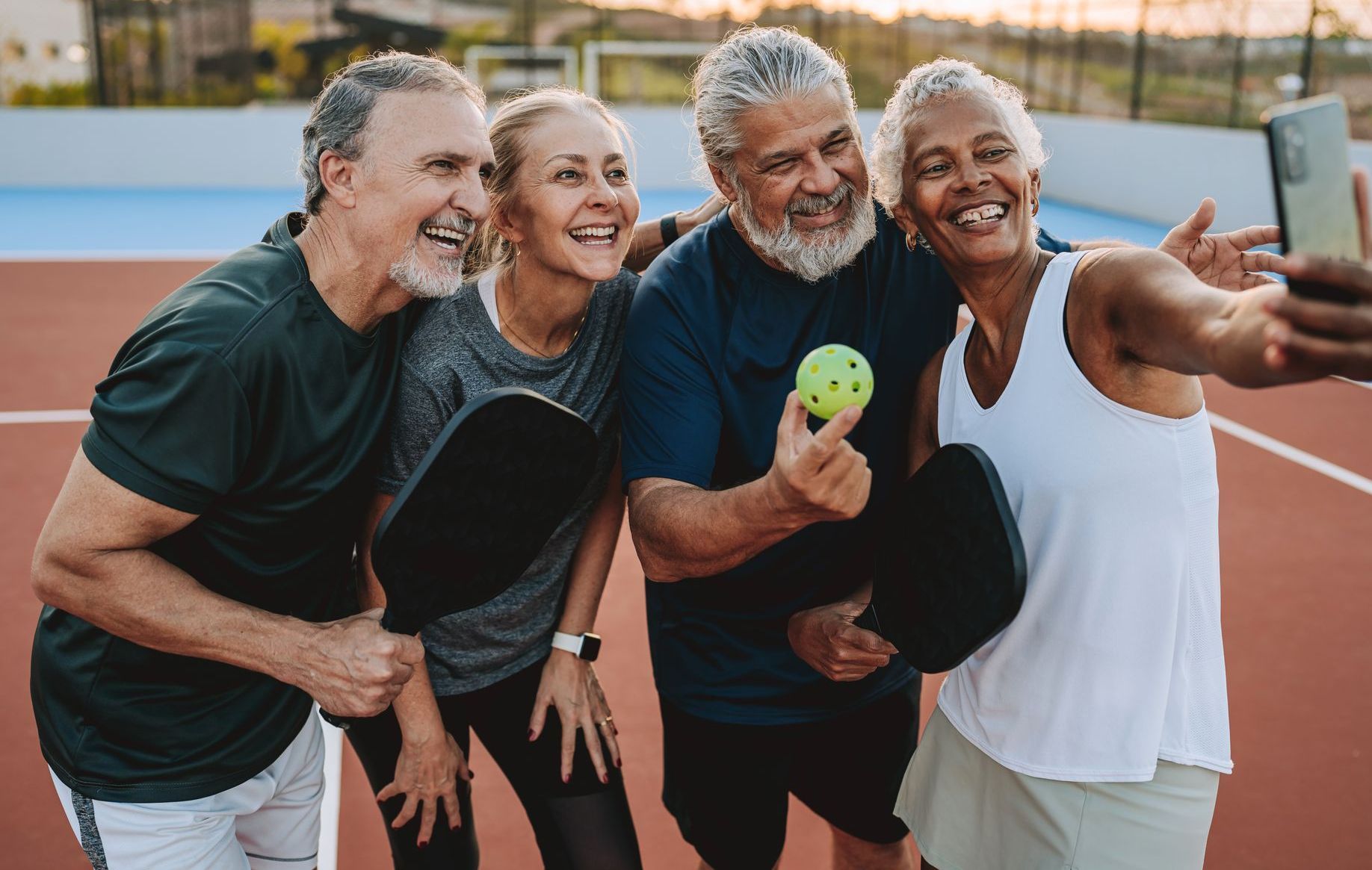 Four smiling people take a selfie on a pickleball court, holding paddles and ball.
