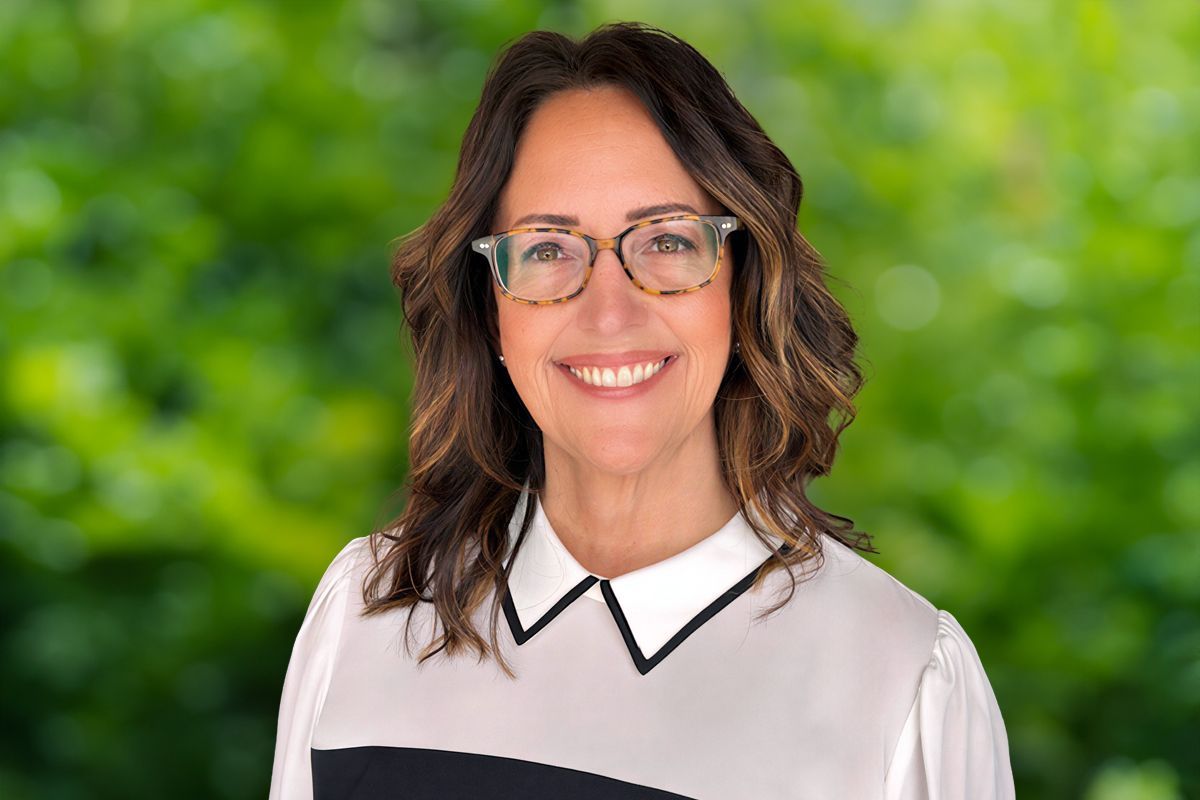 Woman with glasses smiles; wearing white shirt with black accents, set against a blurred green backdrop.