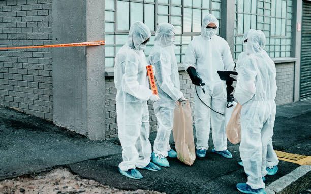 Four people in white protective suits stand outside a building, holding bags and equipment.