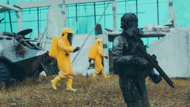 People in yellow hazmat suits and black tactical gear move through a wrecked industrial yard.