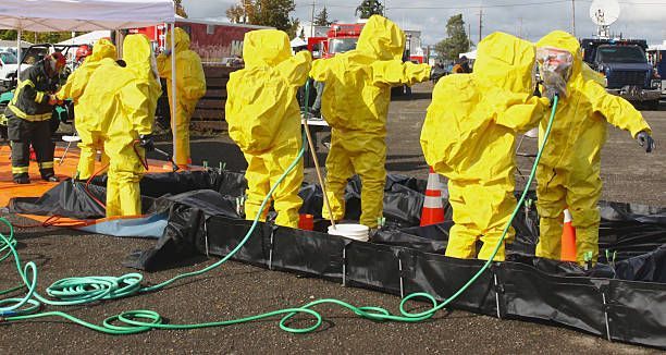 Workers in yellow hazmat suits handling black debris and hoses at an outdoor cleanup site