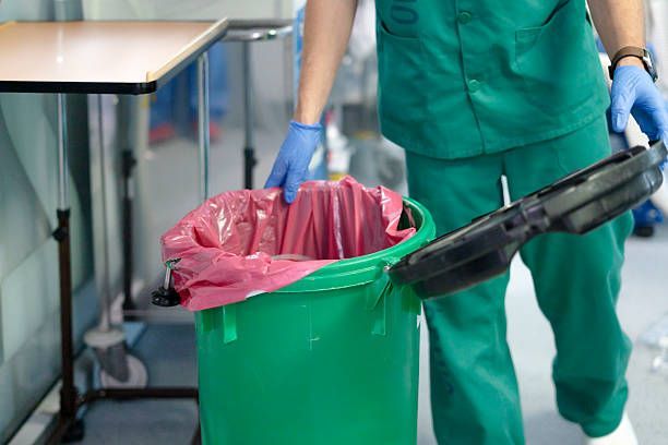 Worker in green scrubs and blue gloves empties a black trash bag into a green bin.