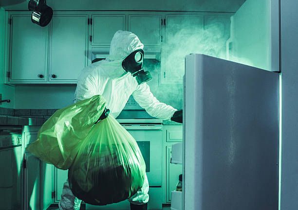 Person in hazmat suit reaching into an open refrigerator in a green-lit kitchen.