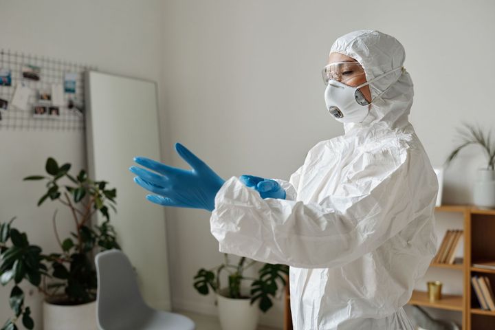 Person in white protective suit and blue gloves standing in a room, adjusting gloves near a mirror and plants