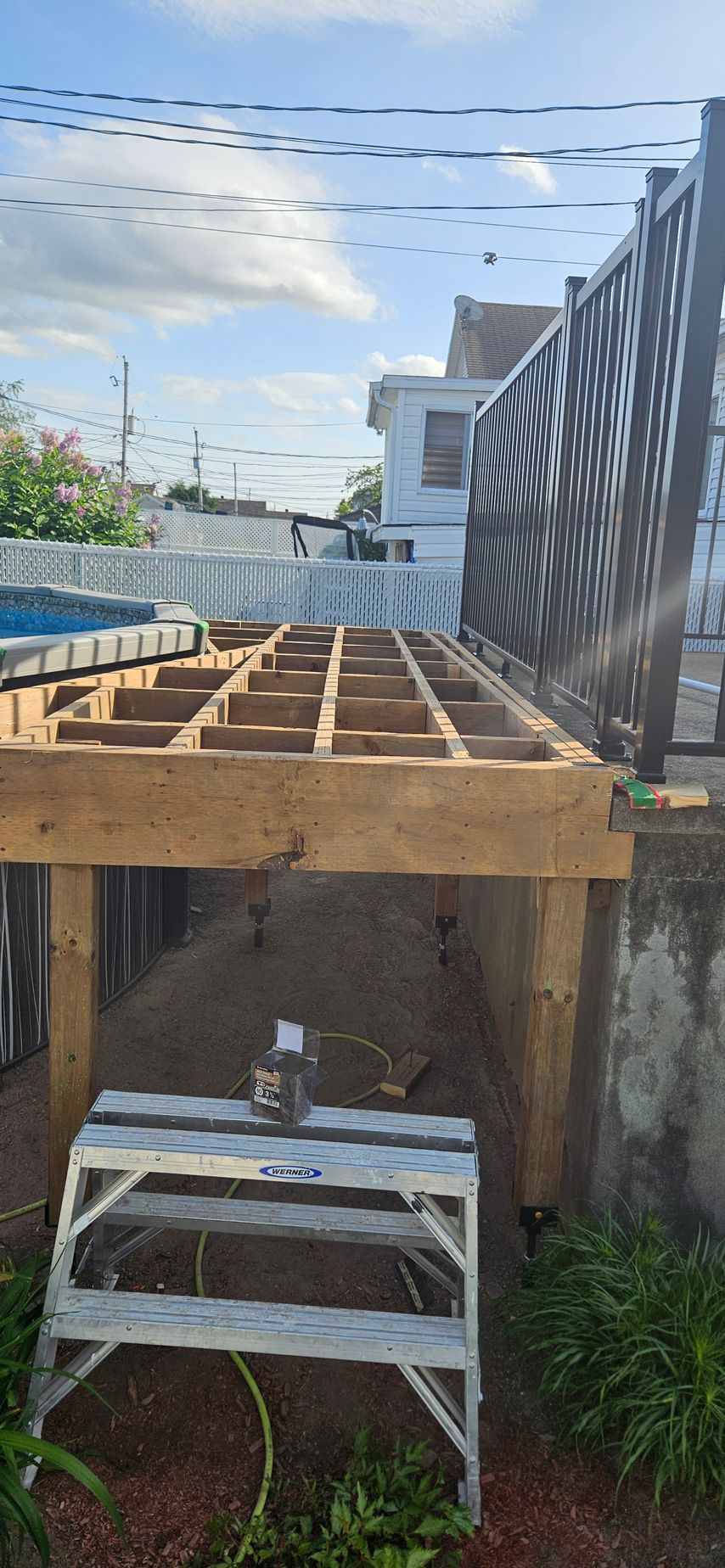 Une charpente de terrasse en construction avec un escabeau en aluminium en dessous. Le ciel est bleu avec quelques nuages.