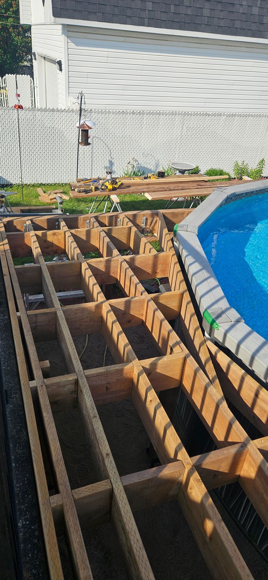 Chantier de construction avec terrasse en bois apparente adjacente à une piscine. Une clôture blanche et un bâtiment sont visibles en arrière-plan.