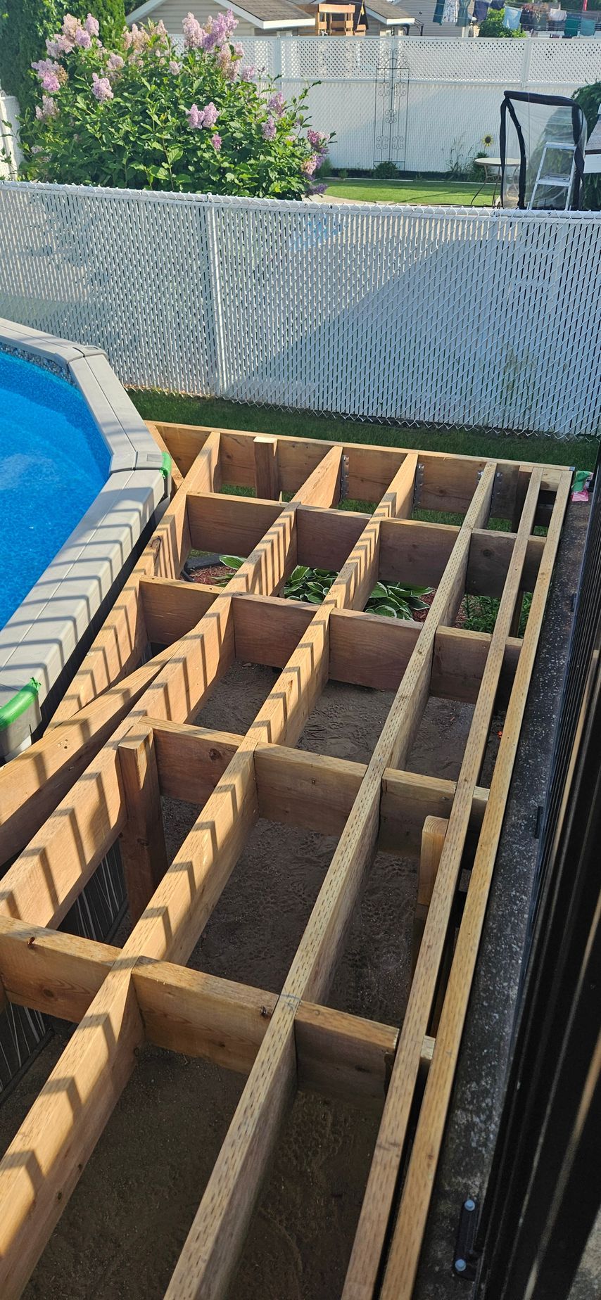 Une terrasse en construction à côté d'une piscine. La structure est en bois, avec une clôture et de la verdure en arrière-plan.