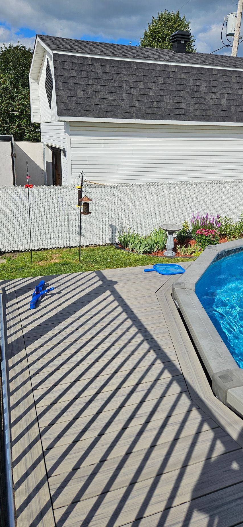 Une cour arrière avec une piscine, une terrasse en bois, une clôture blanche et un bâtiment blanc avec un toit gris foncé.