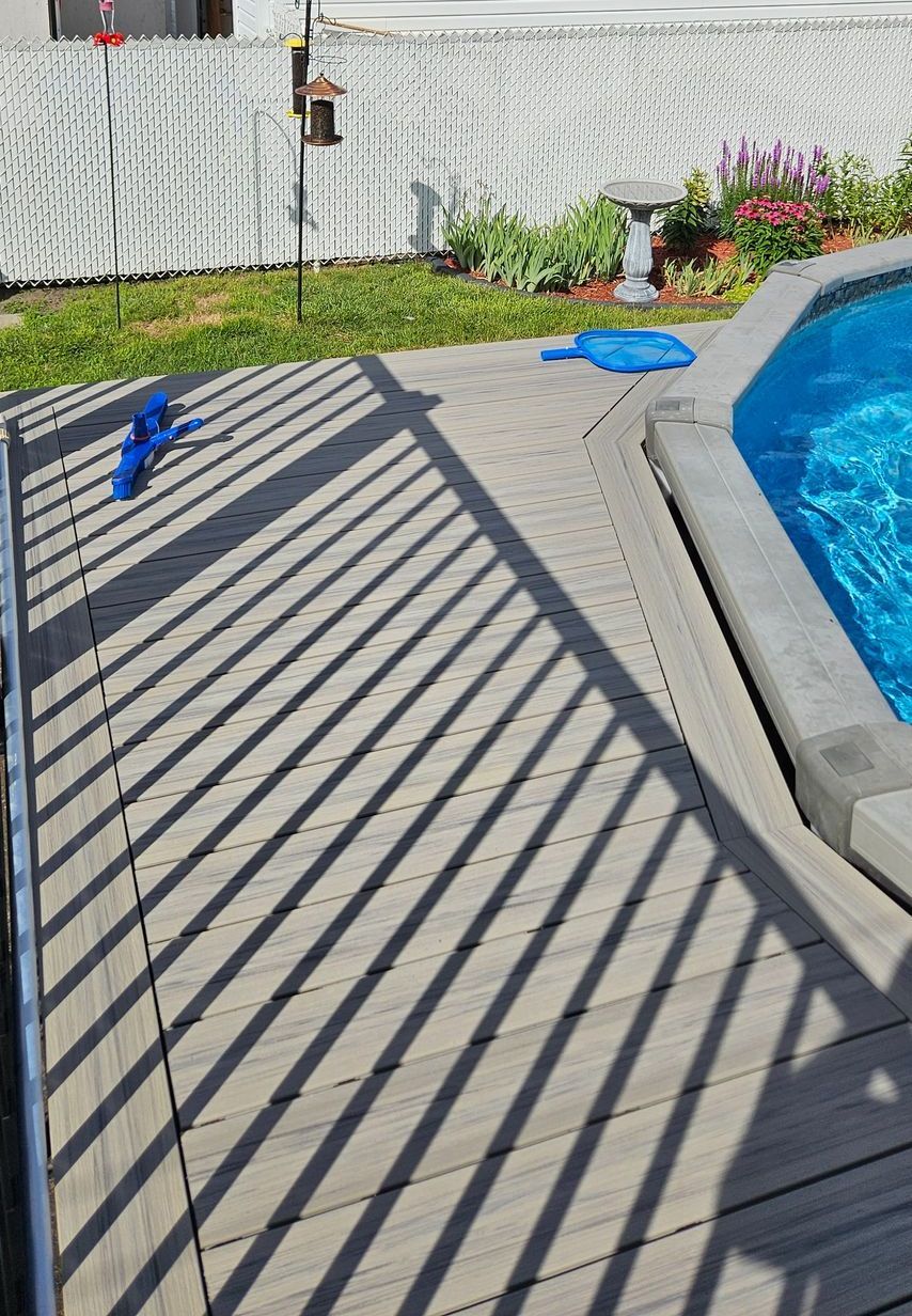 Terrasse en bois avec balustrade projetant des ombres, à côté d'une piscine. Une bâche bleue et des fleurs sont visibles en arrière-plan.