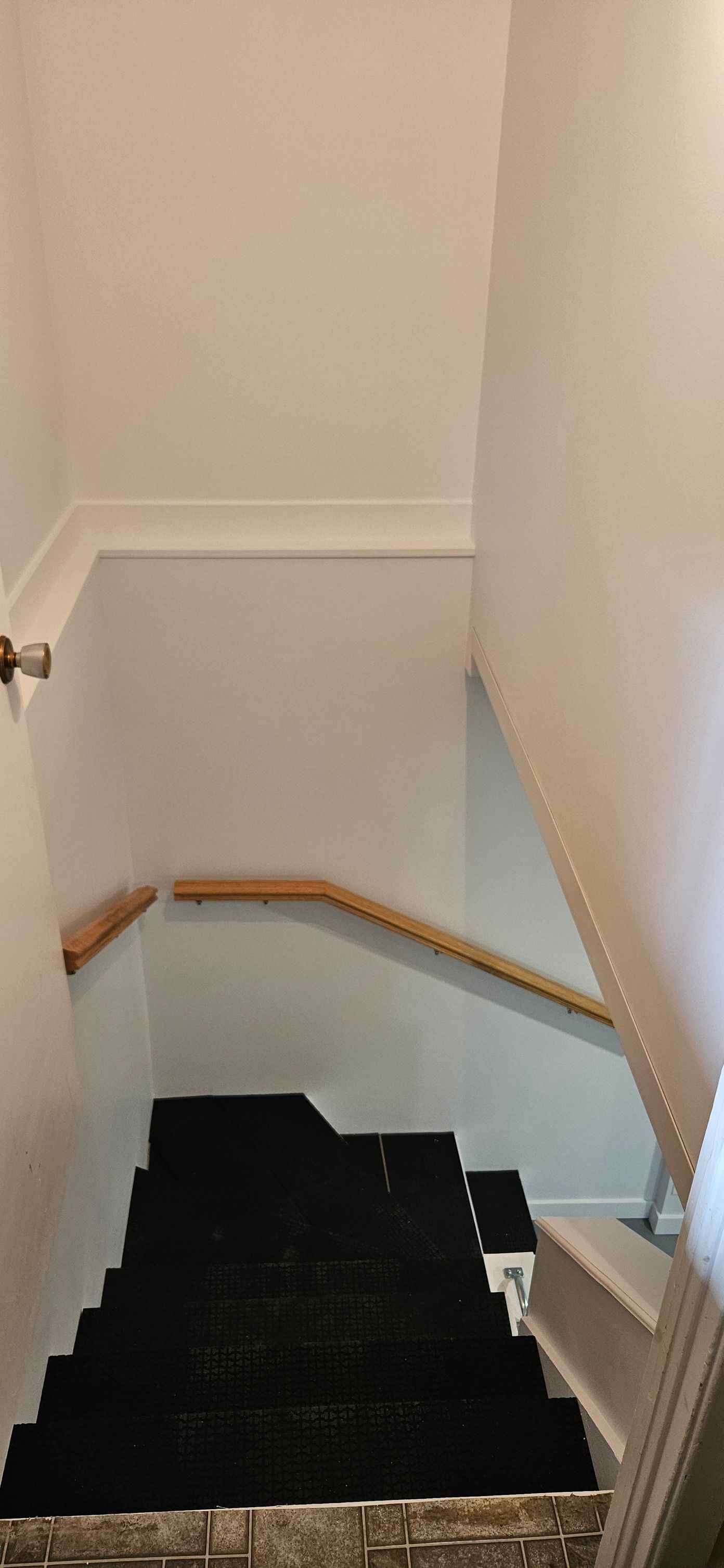 Un escalier menant vers le bas, avec des marches recouvertes de moquette noire, des rampes en bois et des murs blancs.