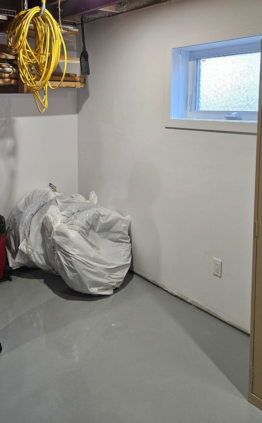Un coin de sous-sol avec une petite fenêtre, un grand sac recouvert de plastique, un sol gris et des murs blancs. Un cordon jaune pend du plafond.