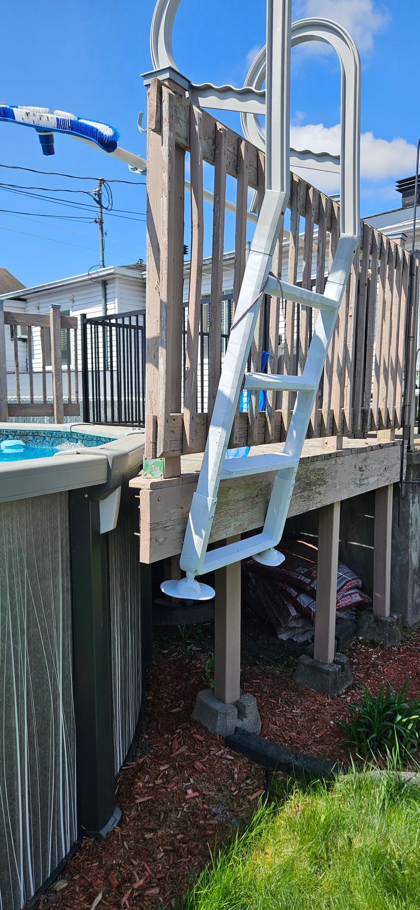 Vue depuis la piscine : une échelle blanche fixée à une terrasse en bois mène à une piscine hors-sol. Ciel bleu visible en arrière-plan.
