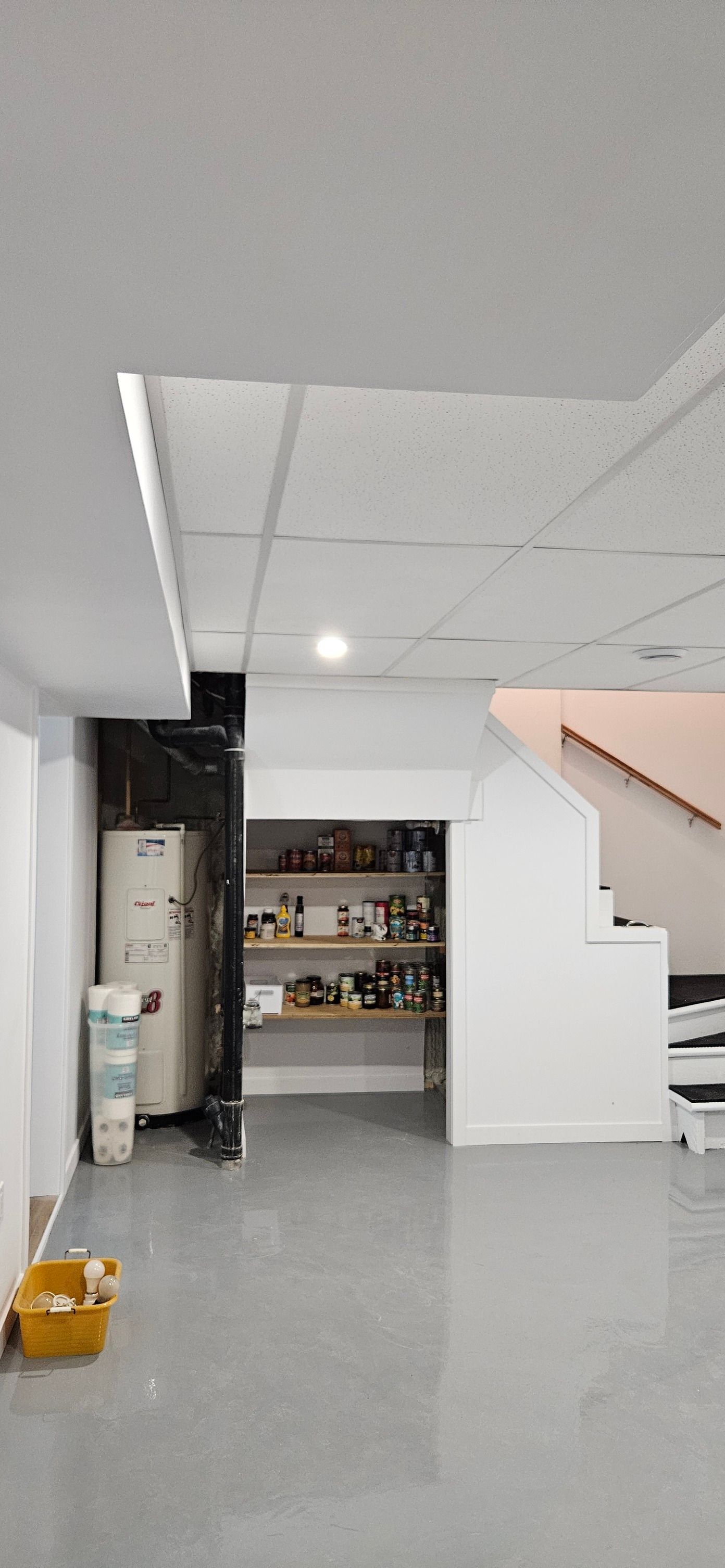 Sous-sol intérieur avec garde-manger et escalier. Sol gris et murs blancs avec étagères remplies de marchandises visibles.