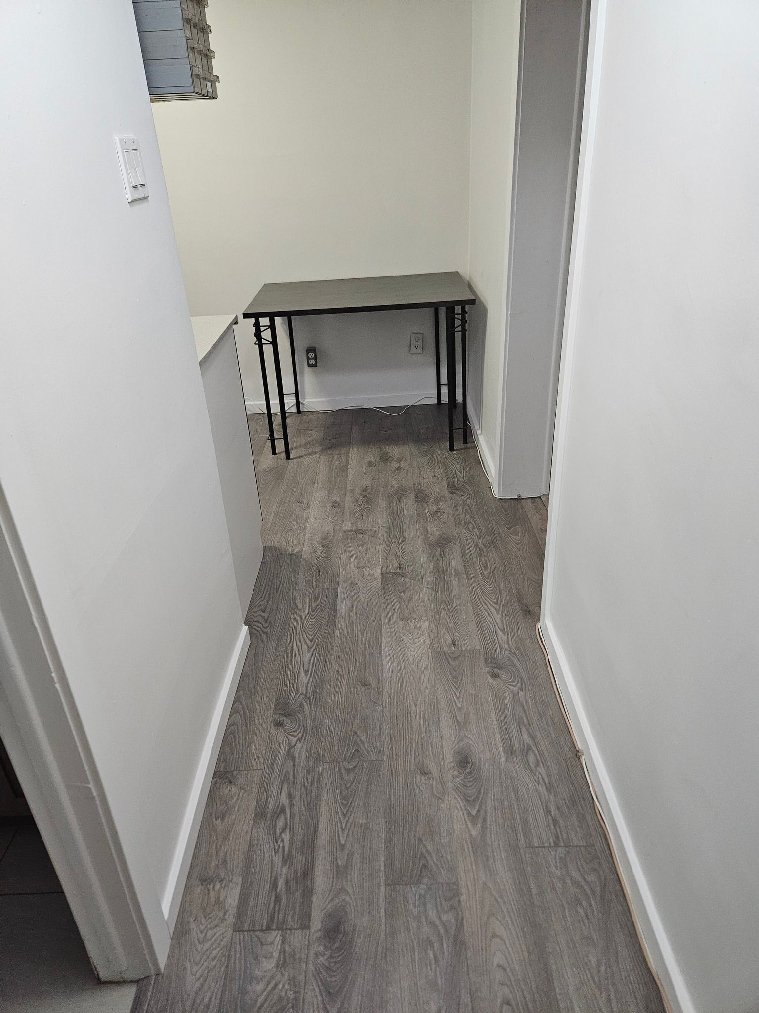 Couloir étroit avec parquet imitation bois gris menant à un petit bureau. Des murs blancs encadrent l'espace.