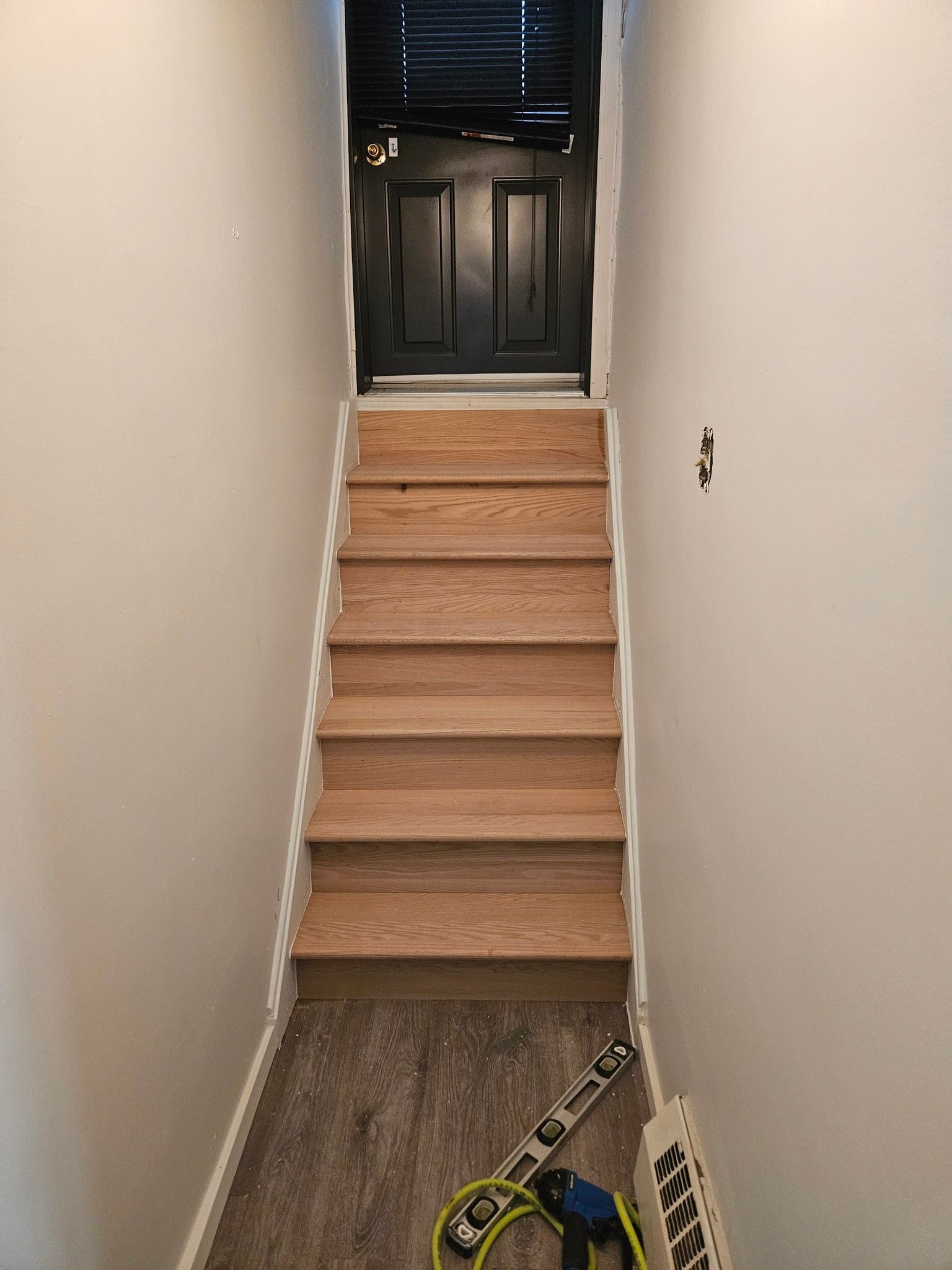 Escalier menant à une porte noire. Marches en bois clair avec contremarches blanches et murs blancs.