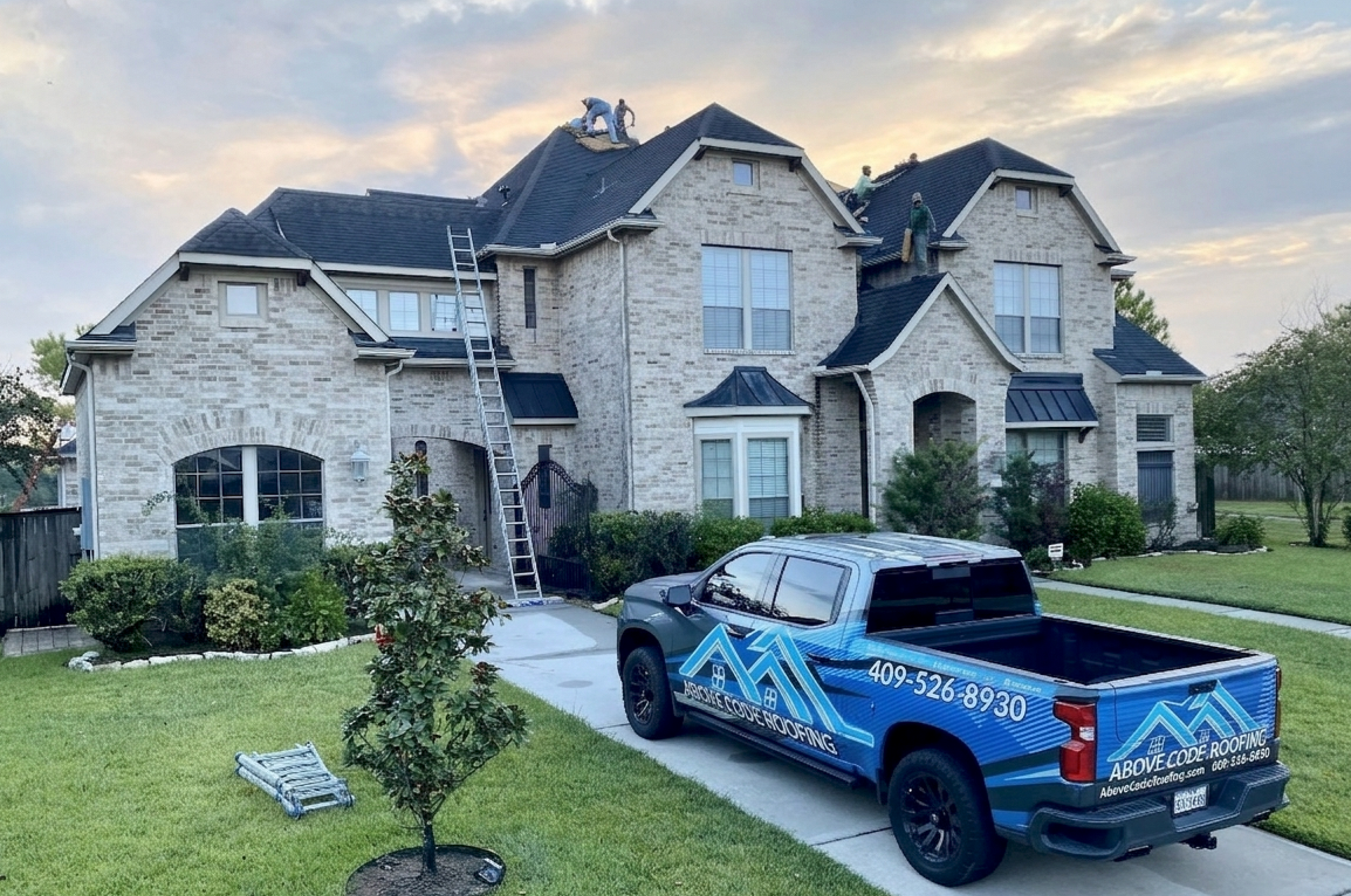 A blue company truck is parked in the driveway of a two-story brick house, where workers are installing a new roof.