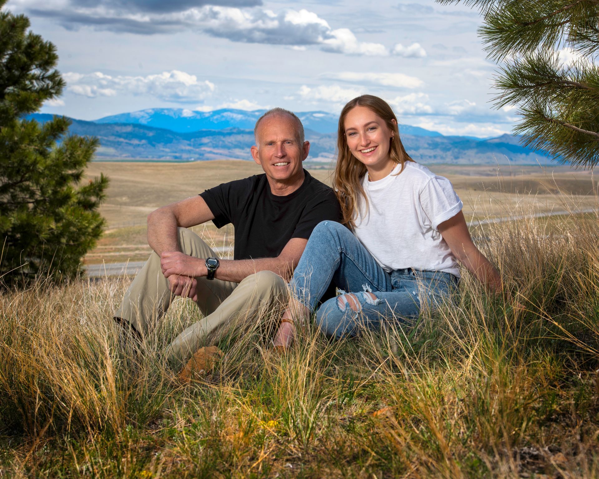A man and a woman are sitting in the grass with mountains in the background.