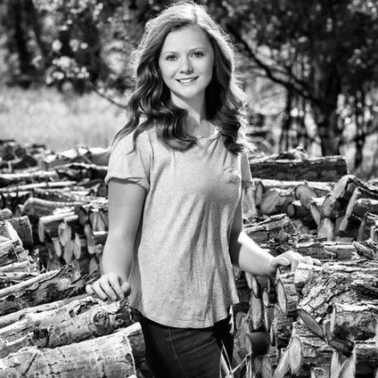 A young woman is standing in front of a pile of logs.