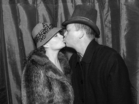 A man and a woman are kissing in front of a curtain . the woman is wearing a fur coat and hat.