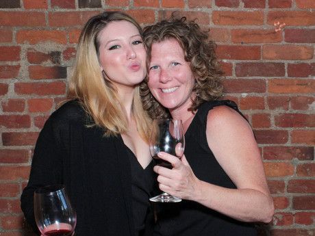 Two women are posing for a picture with wine glasses in front of a brick wall.