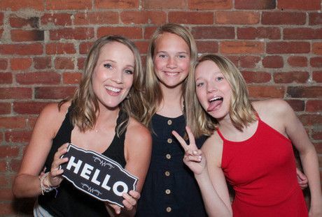 Three girls are posing for a picture in front of a brick wall while holding a hello sign.