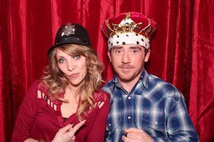 A man and a woman are posing for a picture in a photo booth . the man is wearing a crown.