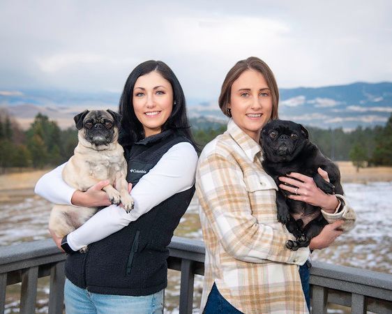 Two women are holding two pug dogs on a bridge.