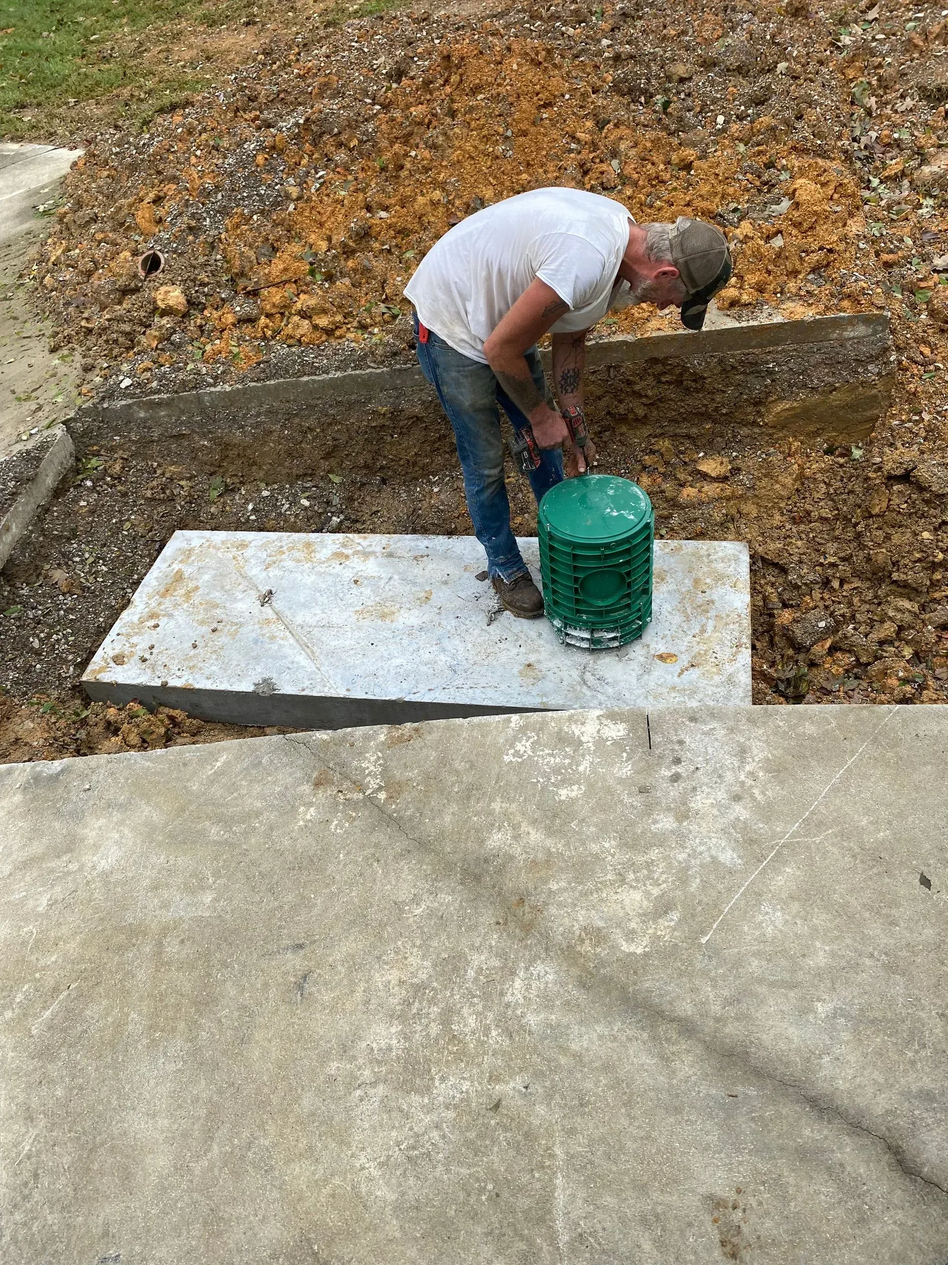 A man is kneeling down on a concrete slab next to a green bucket.