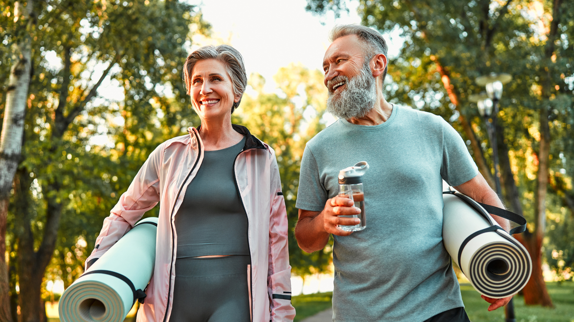 A man and a woman are walking in a park holding yoga mats.