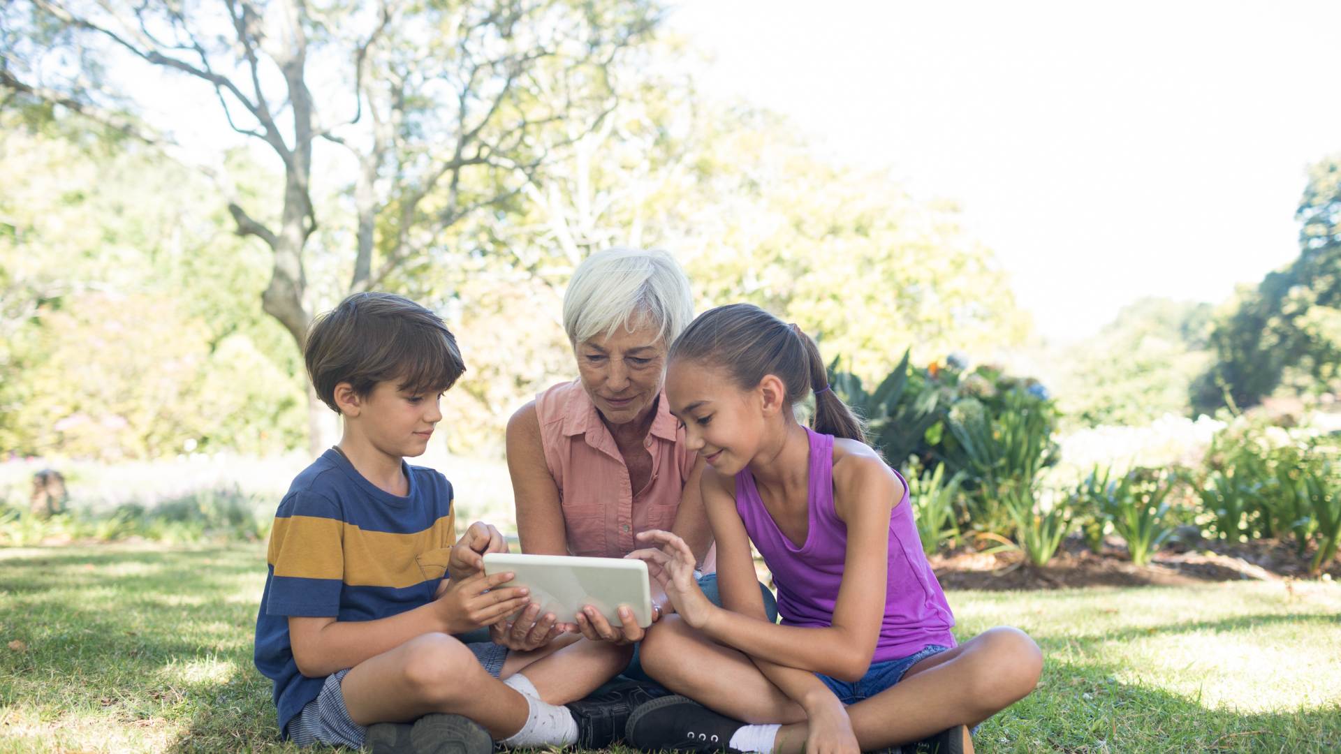 A woman and two children are sitting on the grass looking at a tablet.