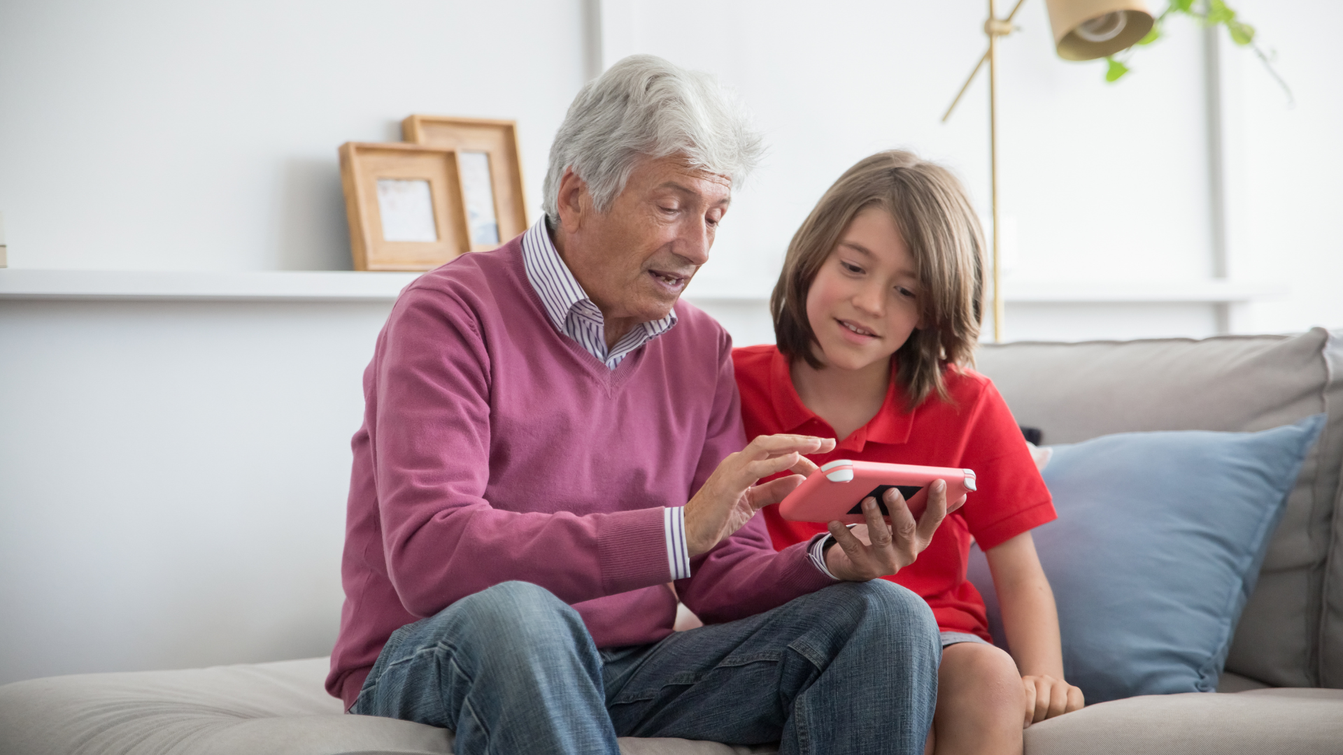 An elderly man and a young boy are sitting on a couch looking at a tablet.