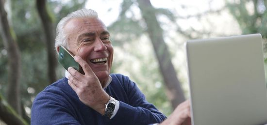 An elderly man is talking on a cell phone while using a laptop computer.