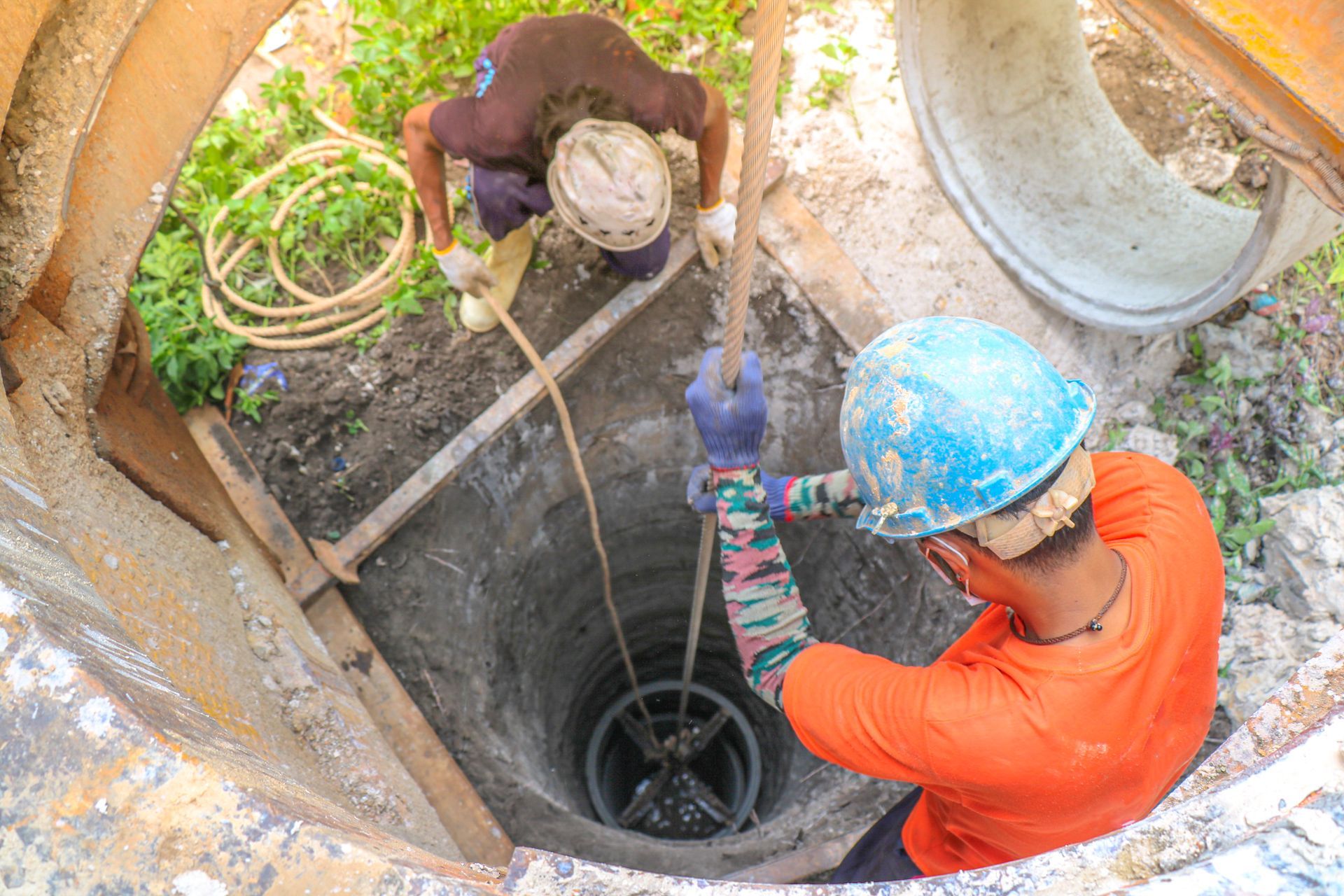 Two workers lowering equipment into a well. One in orange shirt, other in dark shirt; both wearing hard hats.