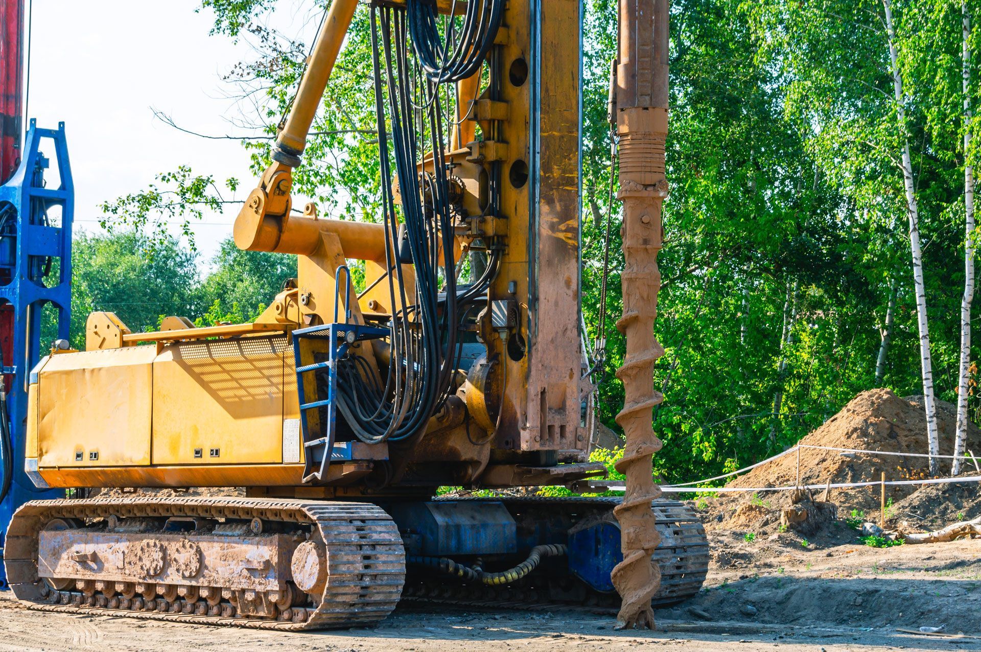 Yellow drilling rig on tracks, next to a pile of dirt, forest in the background.